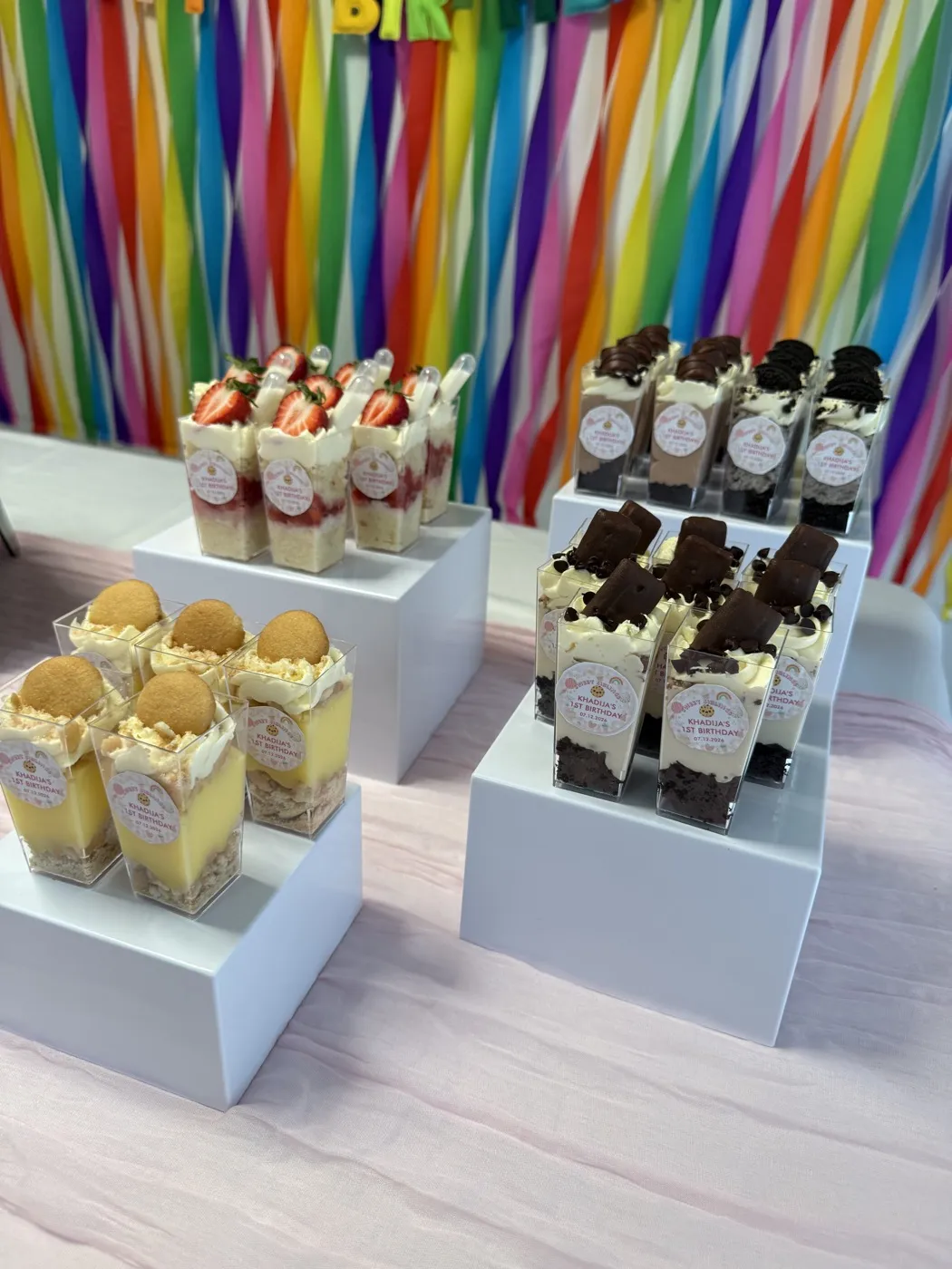 Vanilla mousse, strawberry tres leches, brownie cheesecake, and oreo dessert cups on white display risers at a first birthday